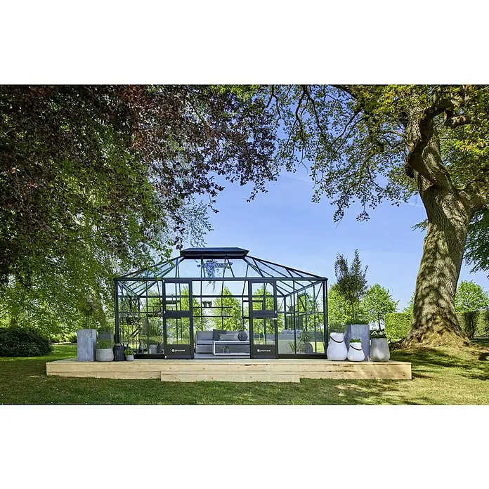 Juliana Gewächshaus Grand Oase - Außenmaß inkl. Dachüberstand (B x T): 368 x 511 cm, 3 mm, Anthrazit/Schwarz - Ein Juliana Gewächshaus mit dunklem Rahmen steht auf einer Holzplattform im Garten, umgeben von Bäumen und Pflanzen.