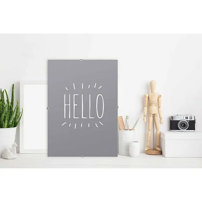 Rahmenloser Bilderrahmen - 70 x 100 cm, Normalglas - Graues Poster mit dem weißen Schriftzug "HELLO", umgeben von Deko-Elementen wie Pflanze, Kamera und Holzpuppe.