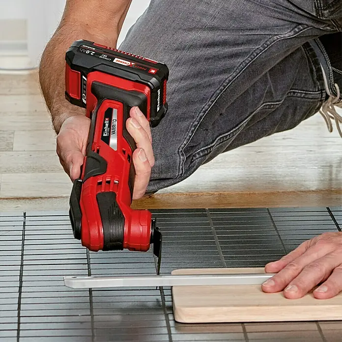 Einhell PXC 18V Multiherramienta de batería Varrito - 18 V, Sin batería - Un hombre usa una multiherramienta roja Einhell VARRITO para cortar un trozo de madera sobre una superficie cuadriculada.