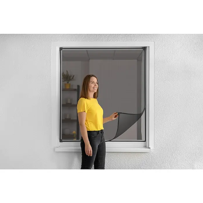 Schellenberg Insect Stop Mosquitera Flex - An x Al: 120 x 120 cm, Montaje de la mosquitera: Fijación magnética, Color bastidor: Antracita - Mujer abriendo una ventana con una mosquitera oscura. La pantalla es oscura y la ventana está enmarcada en blanco.