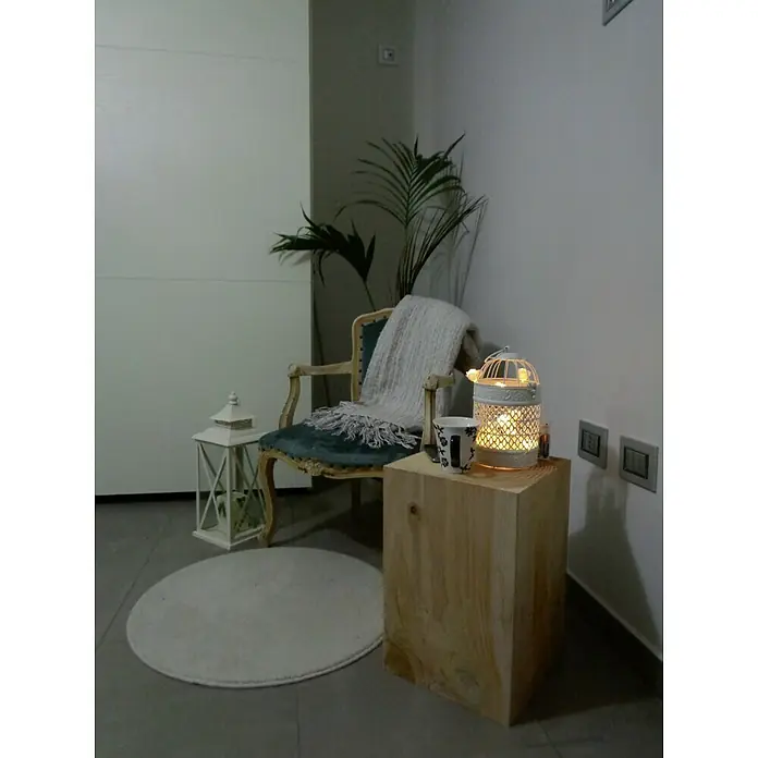 Bloque de madera maciza cuadrado - Abeto, L x An x Al: 30 x 35 x 45 cm - La imagen muestra un rincón acogedor con una silla, una mesa auxiliar con una lámpara y una planta, creando un ambiente relajante.
