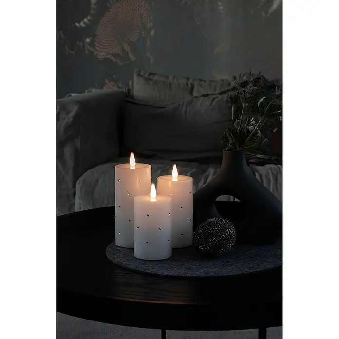 Konstsmide LED-Kerze - Weiß, Höhe: 11,8 cm, 7,5 cm, Wachs - Nahaufnahme von drei weißen LED-Kerzen mit silbernen Akzenten, die auf einem runden, dunklen Tisch mit einer eleganten Vase und Dekoration stehen.
