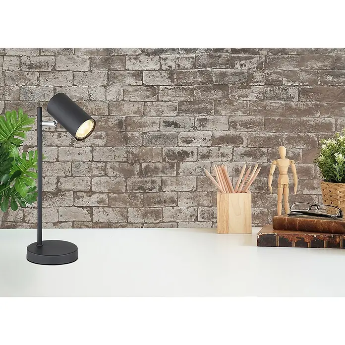 Globo Lámpara de sobremesa Robby - Ø x Al: 12 x 35 cm, GU10 - Un primer plano de una lámpara de escritorio negra con un cabezal ajustable que emite una luz brillante, colocada sobre un escritorio blanco contra una pared de ladrillo.