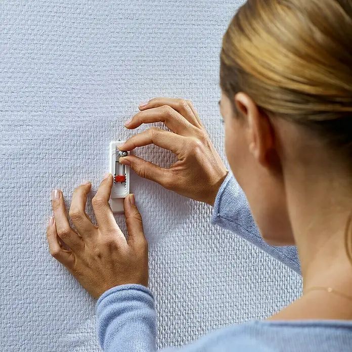 Tesa Verstellbare Klebeschraube - Geeignet für: Tapeten, Belastbarkeit: 1 kg, 2 Stk., Weiß - Eine Frau befestigt ein weißes, längliches Gerät mit rotem Detail an einer weißen Wand, um eine sichere Aufhängelösung zu schaffen.