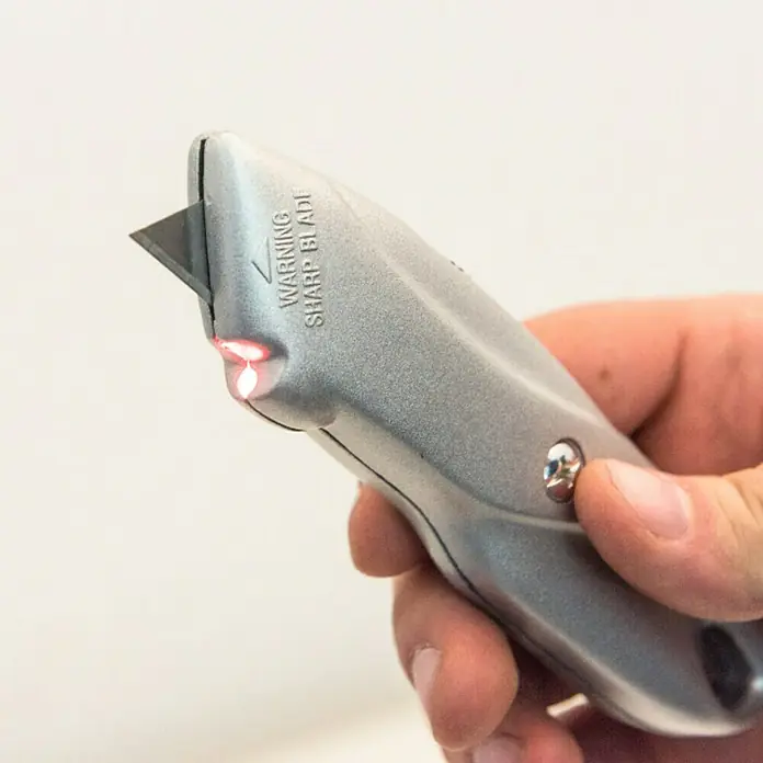 Connex Cuchillo cúter - Metal, Plateado - Primer plano de un cúter gris con una hoja afilada, sostenido por una mano. Se puede ver un mensaje de advertencia en el cuerpo.
