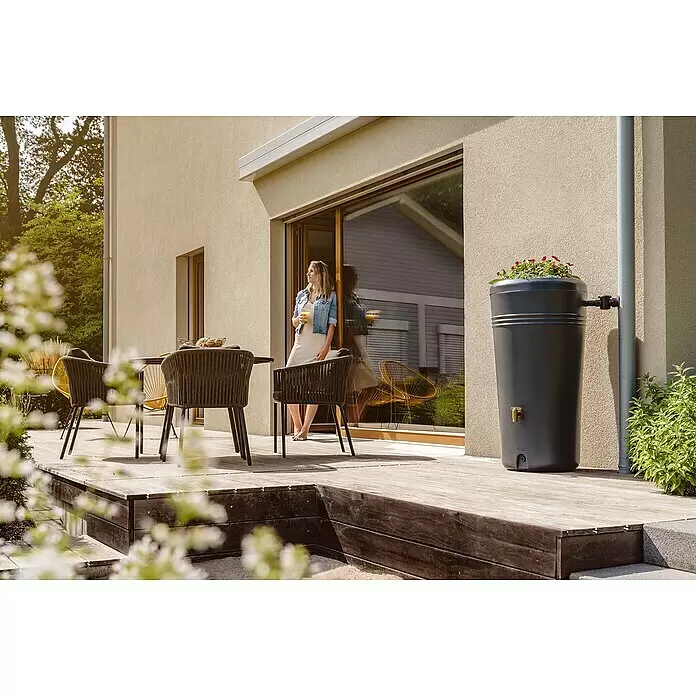 Garantia Regenspeicher Azura 2in1 - 230 l, Graphite - Dunkelgraues Kunststoff-Regenfass sammelt Regenwasser vom Fallrohr auf Holzterrasse, bepflanzt mit roten Blumen.