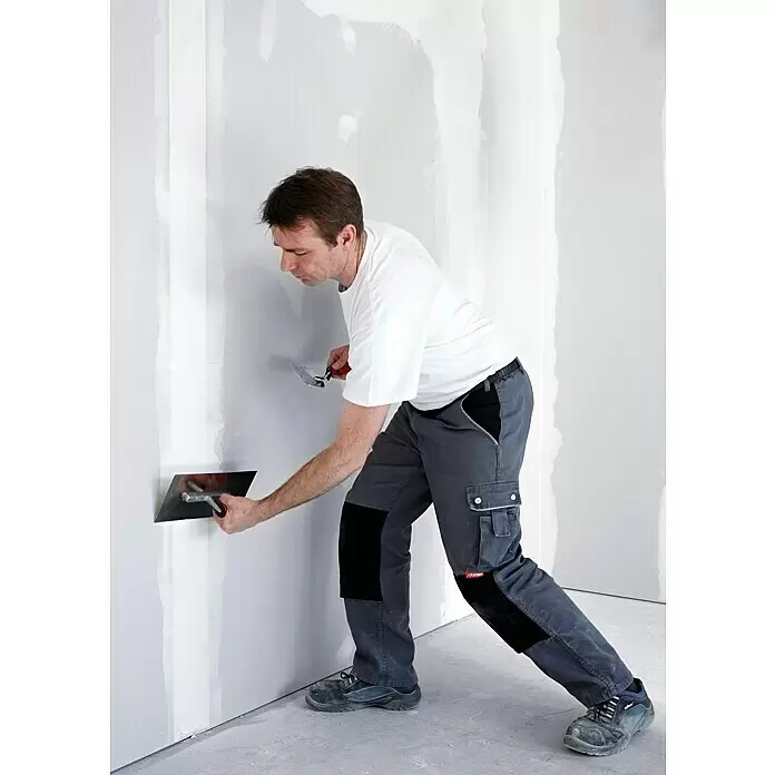 Rigips Flächenspachtel - 5 kg - Arbeiter in dunkler Arbeitshose mit Kniepolstern trägt Spachtelmasse mit einer Glättkelle auf eine Wand auf.