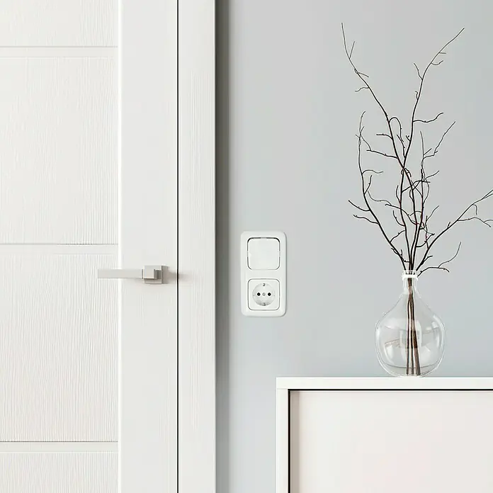 Busch-Jaeger Reflex SI Abdeckrahmen 2512-214 - Alpinweiß, 2-fach, Kunststoff - Nahaufnahme einer weißen Steckdose mit Schalter an einer grauen Wand neben einer weißen Tür und einem minimalistischen Tisch.