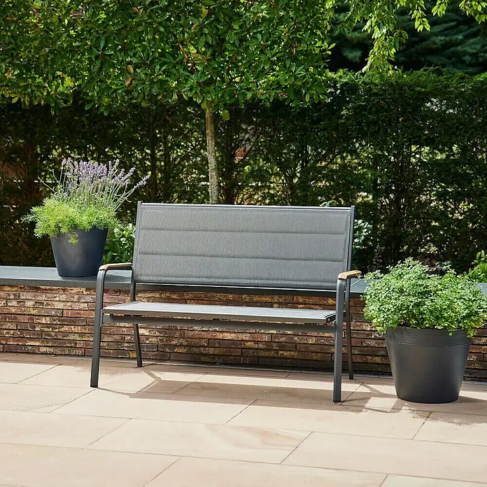 Siena Garden Tesoro Gartenbank - B x T x H: 138,5 x 67 x 97 cm, Anthrazit, Stapelbar, Teak Armlehnen - Eine graue Gartenbank mit Holzarmlehnen steht auf einer Terrasse, flankiert von Blumentöpfen, die das Ambiente verschönern.