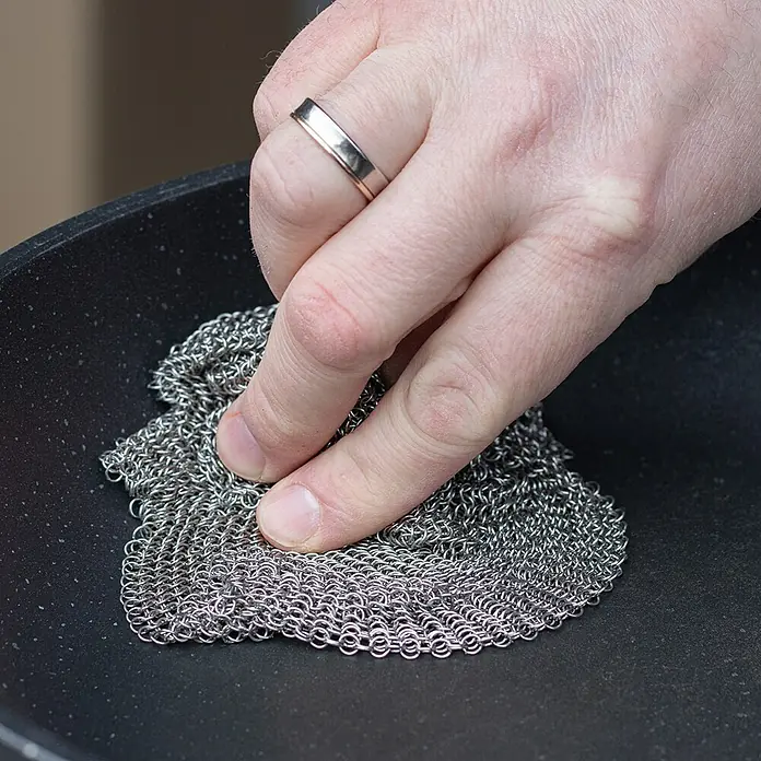 Grillreiniger Ringreiniger - L x B: 20 x 20 cm, Edelstahl - Eine Hand reinigt eine Bratpfanne mit einem Kettengeflecht-Schrubber. Ideal zum schonenden Entfernen von Rückständen ohne die Oberfläche zu beschädigen.