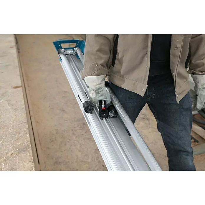 Bosch Professional Transport- & Arbeitstisch GTA 3800 - Arbeitshöhe: 824 mm, Traglast: 250 kg, Geeignet für: Kappsägen - Eine Person trägt eine lange Aluminiumfliesenschneidemaschine in einer Werkstatt. Das Gerät ist blau und silberfarben.
