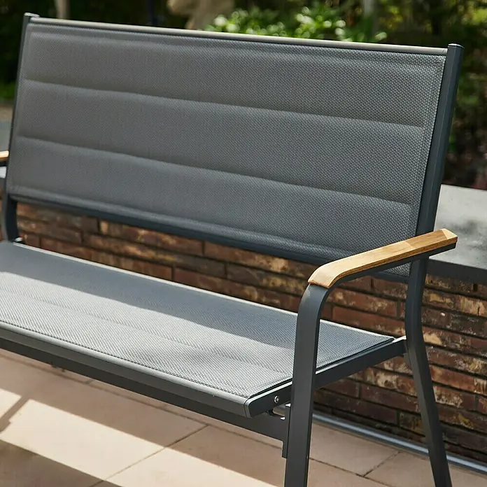 Siena Garden Tesoro Gartenbank - B x T x H: 138,5 x 67 x 97 cm, Anthrazit, Stapelbar, Teak Armlehnen - Nahaufnahme einer grauen Gartenbank mit Holzarmlehnen vor einer Ziegelmauer.