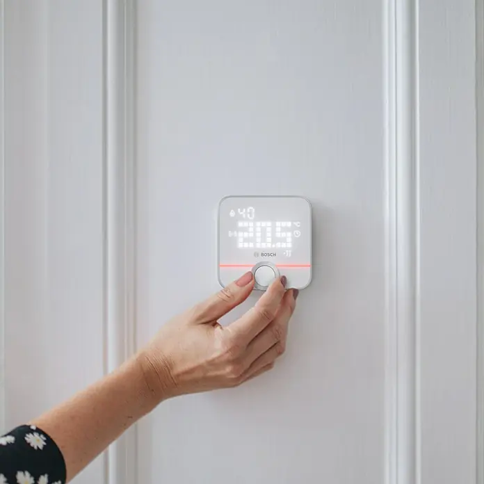 Bosch Smart Home Raumthermostat II - Weiß, Silber, L x B x H: 87 x 86 x 35 mm - Eine Hand bedient einen smarten Bosch Thermostat an einer weißen Wand, der die Temperatur auf 20.5 Grad Celsius einstellt.