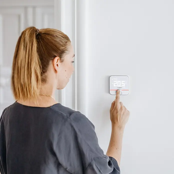 Bosch Smart Home Raumthermostat II Fußbodenheizung - 230 V, LED-Display, L x B x H: 35 x 86 x 87 mm - Eine Frau bedient einen Bosch Smart Home Thermostat an einer weißen Wand, um die Temperatur im Raum zu regulieren.