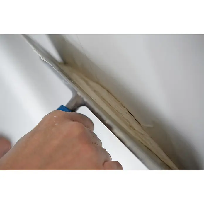 Rigips Fugenspachtel ProMix Plus18 kg, Gebrauchsfertig - Nahaufnahme einer Hand, die mit einer Glättkelle Spachtelmasse auf eine weiße Wand aufträgt, um eine glatte Oberfläche zu erzielen.