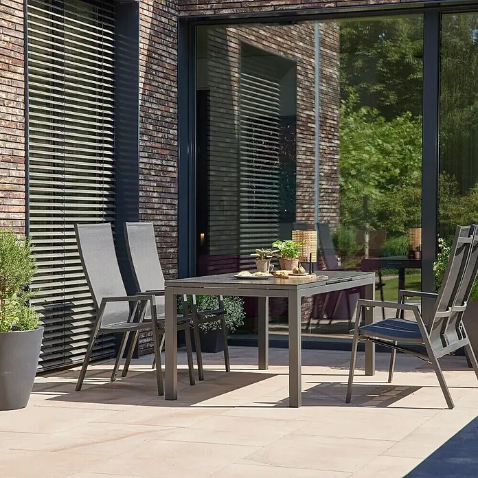 Siena Garden Stapelstuhl Savona - Aluminium, Stapelbar, Anthrazit, B x T x H: 57,5 x 64 x 112,5 cm - Eine graue Esstischgarnitur mit vier Stühlen aus Metall steht auf einer Terrasse vor einer Backsteinmauer. Ideal für den Garten.