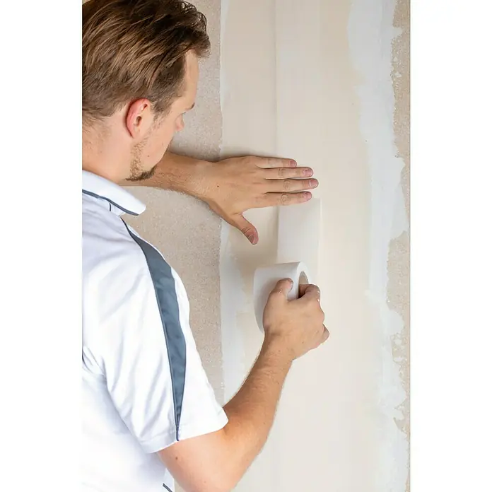 Rigips Rigidur Glasgewebeband - 50 m x 70 mm - Ein Handwerker tapeziert eine Wand mit einer Rolle Malerkrepp, um eine saubere Linie zu ziehen. Das Malerkrepp schützt die bereits fertiggestellte Fläche.