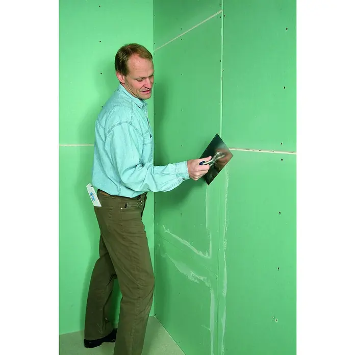 Rigips VARIO Fugenspachtel - 2,5 kg, Imprägniert - Ein Mann glättet eine grüne Gipskartonwand mit einem Spachtel. Im Hintergrund sind Schraubenlöcher und Fugen erkennbar.