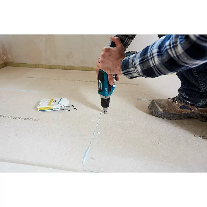 Rigips Rigidur Estrichelement - 1.500 x 500 x 25 mm - Eine Person verwendet einen Makita-Bohrer, um helle Bodenplatten zu verschrauben. Eine Schachtel Rigips-Schrauben liegt daneben.