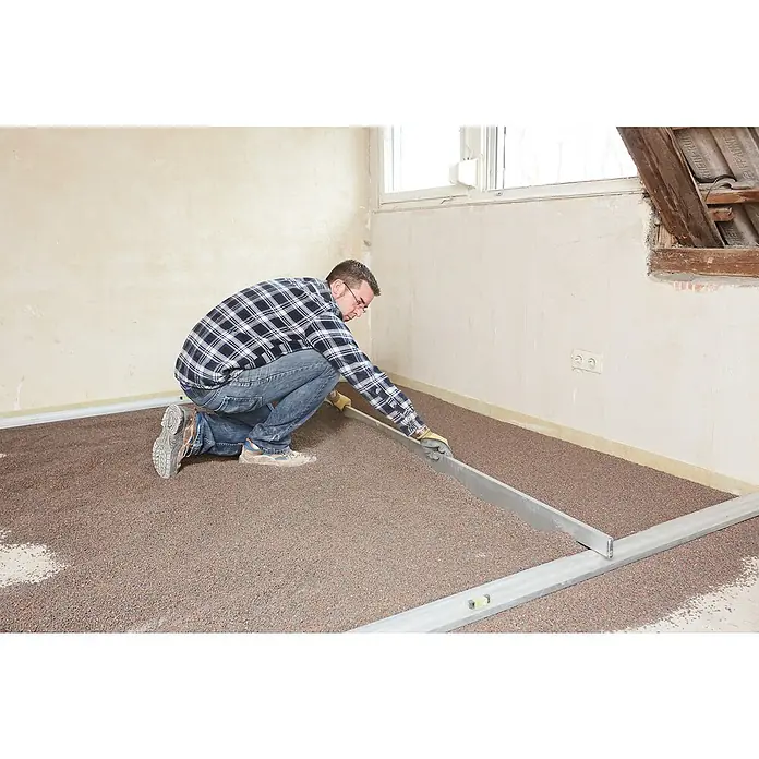 Rigips Rigidur Ausgleichsschüttung - 50 l - Mann verlegt Dämmmaterial auf einem Dachboden. Das Dämmmaterial ist feinkörnig und wird mit einer langen Metallschiene verteilt.