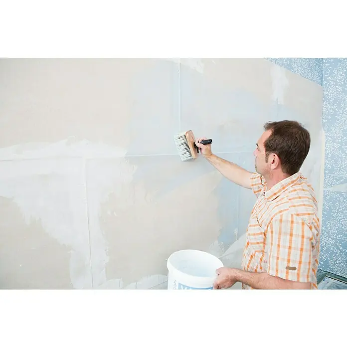 Rigips VARIO Sicherheitsgrundierung - 5 l - Ein Mann trägt mit einem breiten Pinsel Kleister auf eine Wand auf, um Tapeten anzubringen. Er hält einen Eimer Kleister in der Hand.