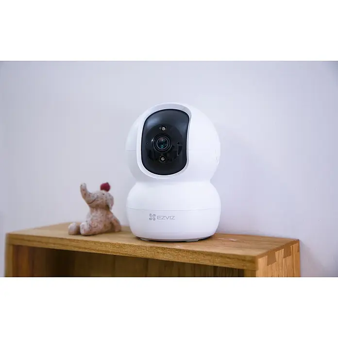 EZVIZ Cámara de vigilancia TY2 con cobertura visual de 360° - Blanco, Ángulo de detección: 360 °, Alcance de detección: 10 m - Primer plano de una cámara de seguridad EZVIZ blanca en un estante de madera con un pequeño elefante. La cámara tiene una lente negra.
