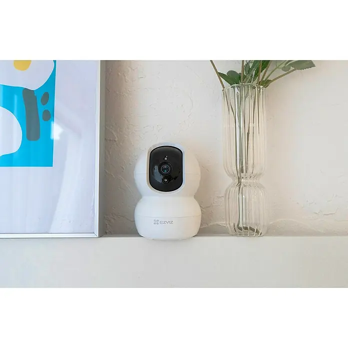 EZVIZ Cámara de vigilancia TY2 con cobertura visual de 360° - Blanco, Ángulo de detección: 360 °, Alcance de detección: 10 m - Primer plano de una cámara de seguridad EZVIZ colocada en un estante junto a un cuadro enmarcado y un jarrón de flores.