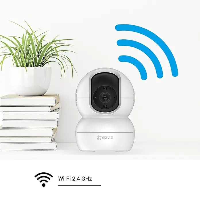 EZVIZ Cámara de vigilancia TY2 con cobertura visual de 360° - Blanco, Ángulo de detección: 360 °, Alcance de detección: 10 m - La imagen muestra una cámara de seguridad EZVIZ con un símbolo de Wi-Fi, junto a una maceta con plantas verdes sobre una pila de libros.