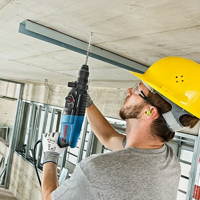 Bosch Professional Martillo combinado GBH 2-26 F - 830 W, 2,7 J, Alojamiento SDS-Plus - Un hombre perforando un techo de concreto con un martillo rotatorio Bosch, usando equipo de seguridad como casco y gafas.