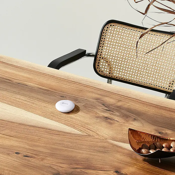 EZVIZ Pack de seguridad Smart Home A3 - 4 pzs., ZigBee 3.0 - Un sensor inteligente redondo se encuentra sobre una mesa de madera junto a una silla. El sensor es compacto y discreto.