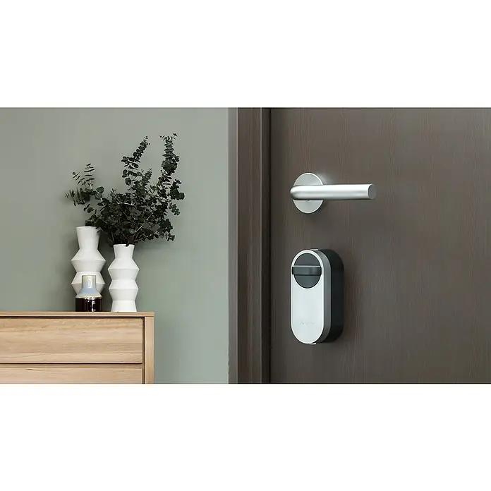 EZVIZ Cerradura electrónica DL01S - Apto para: Puerta, 4 baterías - Primer plano de una moderna cerradura de puerta con manija en una puerta de madera, junto a un aparador con plantas.