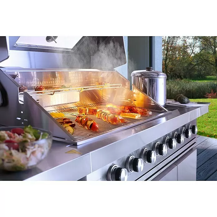 Buschbeck Outdoorküche Bristol - Edelstahl, Silber, Material Grillfläche: Edelstahl, Anzahl Brenner: 6 Stk. - Großer Edelstahl-Gasgrill, auf dem Spieße, Mais und Fleisch im Freien zubereitet werden, mit Topf, Salat und Schneidebrett daneben.