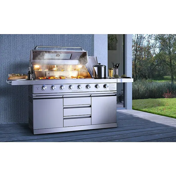 Buschbeck Outdoorküche Bristol - Edelstahl, Silber, Material Grillfläche: Edelstahl, Anzahl Brenner: 6 Stk. - Bild von einem modernen Gasgrill mit Grillgut auf einer Terrasse. Das hochwertige Gerät aus Edelstahl ist mit Bedienknöpfen ausgestattet.