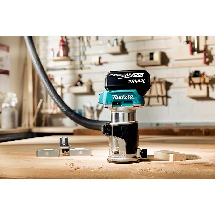 Makita LXT 18V Fresadora multifunción de batería DRT50Z - 18 V, Sin batería, Número de revoluciones de marcha en vacío: 10.000 r.p.m. - 30.000 r.p.m. - Primer plano de una fresadora inalámbrica Makita con una batería conectada a un banco de trabajo, herramientas al fondo.
