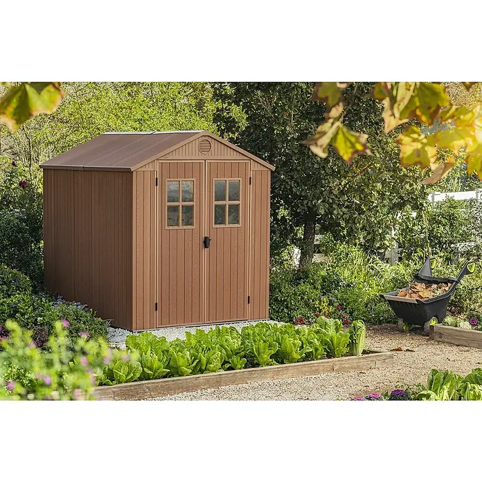 Keter Caseta de jardín Darwin 6X8 II - Medidas ext. (An x Pr): 190 x 244 cm, Plástico, Marrón, 4,5 m² - Una caseta de jardín marrón se encuentra en un jardín con lechos de lechuga. La caseta tiene puertas dobles y un techo a dos aguas con ventilación.