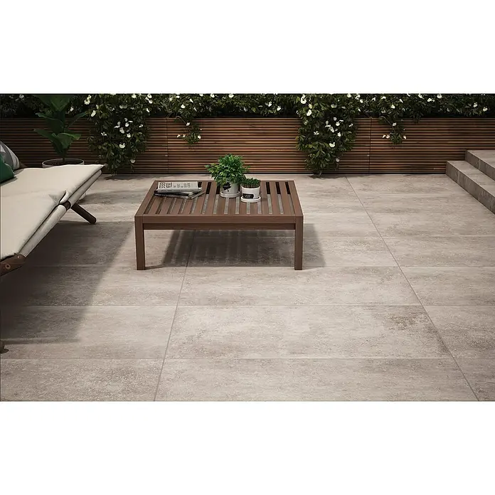 RAK Ceramics Terrassenfliese Volterra 2.0 - 60 x 120 x 2 cm, Sandgrau, Matt - Einladende Terrasse mit grauen Fliesen, einem Holztisch und einem bequemen Liegestuhl, ideal zum Entspannen im Freien.