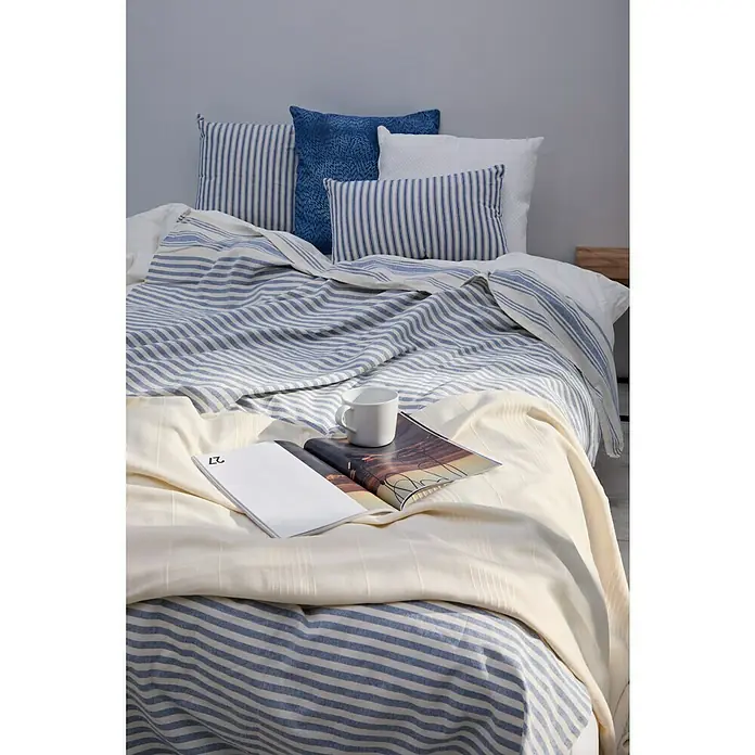 Manta para sofá Ernesta - 150 x 250 cm, Textil, Azul - La imagen muestra una cama acogedora adornada con ropa de cama a rayas azules y blancas, almohadas y una manta ligera, con un libro y una taza.