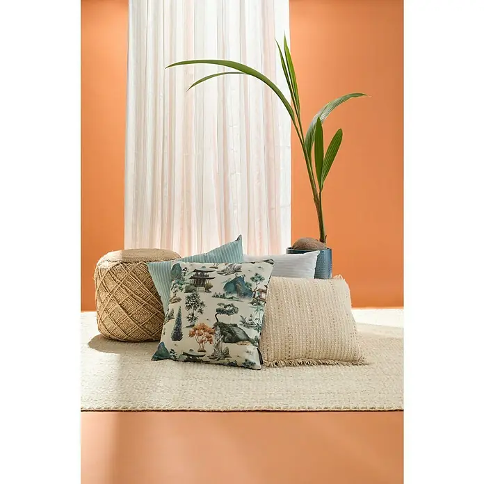 Cojín Dakira - 45 x 45 cm, Textil, Natural, Cuadrado - Ambiente interior con cojines decorativos, un taburete de mimbre y una planta de interior sobre un fondo naranja.