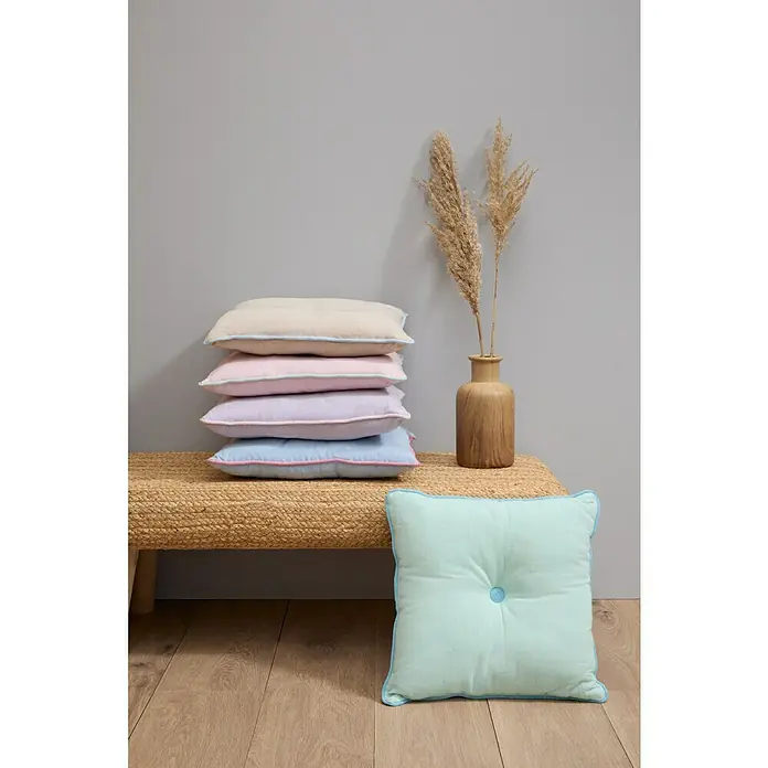 Cojín Botón - 45 x 45 cm, Textil, Lavanda, Cuadrado - Una pila de almohadas de colores con ribetes a juego junto a un jarrón con flores secas en un banco de mimbre, más una almohada adicional.