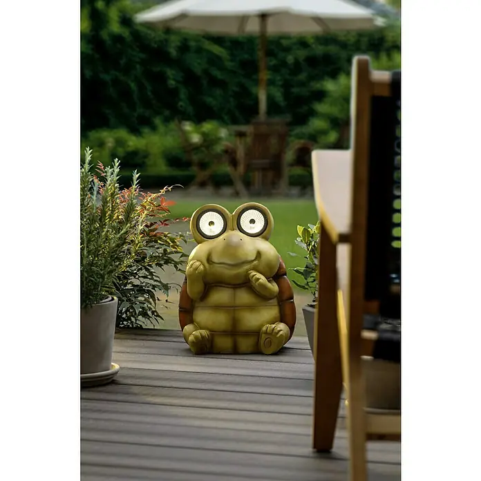 BAUHAUS Solar-Dekoleuchte - Lichtfarbe: Warmweiß, 24 x 27 x 35 cm, Schildkröte - Lächelnde Schildkrötenfigur mit Solaraugen als Dekoration für den Garten, sitzt auf einer Holzterrasse.