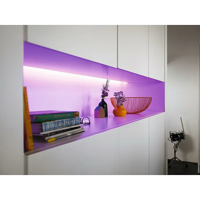 Philips Hue LED-Band Plus Basis - 2 m, RGBW - Einbaufach mit violetter Beleuchtung, das Bücher, dekorative Vasen und eine Schale zeigt und so eine stilvolle Raumatmosphäre schafft.