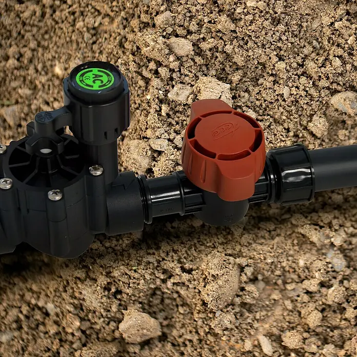 Llave de paso Goteo - 1 ", Polietileno - Primer plano de un sistema de riego con una electroválvula negra y una válvula de corte roja sobre un fondo terroso.