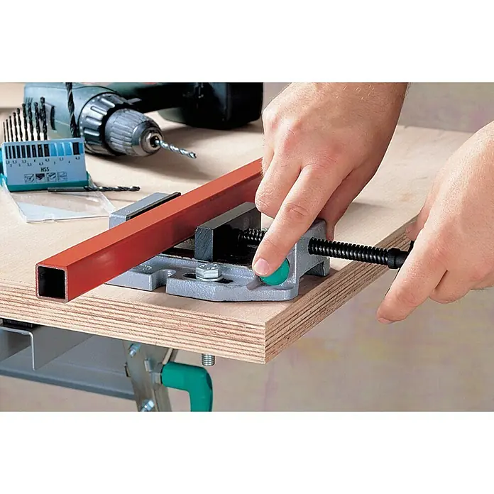 Wolfcraft Mordaza de sujeción - Anchura de sujeción: 80 mm - Un banco de trabajo con un tornillo de banco que sujeta un tubo cuadrado rojo. Al fondo, un taladro inalámbrico y un juego de brocas.