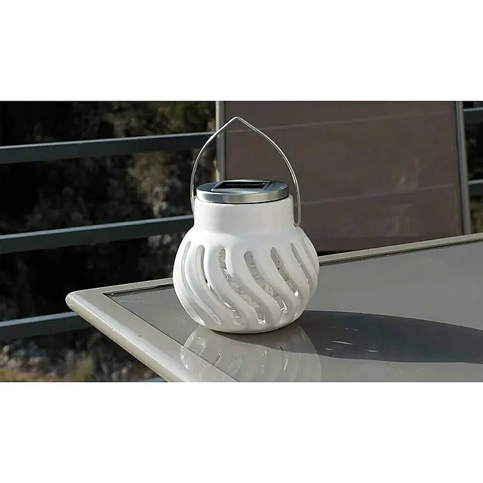 Farolillo antimosquitos solar Oriental - 12 V - Primer plano de una linterna solar de cerámica blanca con asa plateada, colocada sobre una mesa al aire libre.