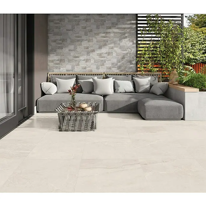 Halcón Pavimento porcelánico Tempo - 60,5 x 60,5 cm, Cotton, Mate, Clase 3 Antideslizante - Un elegante espacio al aire libre con baldosas de suelo beiges, un sofá gris y una mesa de mimbre. Perfecto para relajarse al aire libre.