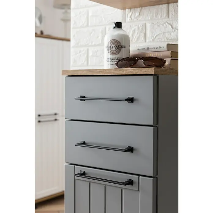 Schildmeyer Jelda Unterschrank - 30,3 x 32,6 x 88,1 cm, Platingrau/Eiche, Matt - Grauer Badezimmerschrank mit drei Schubladen und schwarzen Griffen, gekrönt von einer Holzplatte, auf der Handseife und Sonnenbrille stehen.
