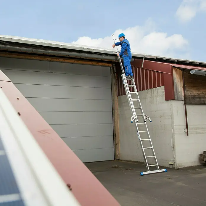 Krause Stabilo Schiebeleiter - Anzahl Tritte: 24 Stk., Arbeitshöhe: 6,85 m, Aluminium - Ein Mann in Arbeitskleidung benutzt eine Leiter, um ein Dach zu reparieren, wobei die robuste Bauweise der Leiter im Vordergrund steht.