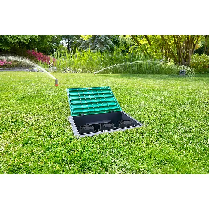 Gardena Sprinklersystem Ventilbox V3 - Passend für: 3 Bewässerungsventile - Eine grüne Bewässerungsanlage mit geöffnetem Deckel ist im Rasen eingebettet, während Sprinkler im Hintergrund den Rasen bewässern.