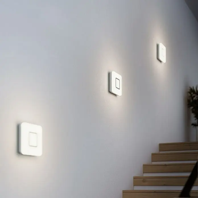 Steinel Sensor-Wand- & Deckenleuchte RS LED M1 S Edelstahl - 8,8 W, L x B x H: 20 x 20 x 5,3 cm, Weiß - Drei quadratische LED-Wandleuchten an einer weißen Wand neben einer Holztreppe. Das Licht ist weich und diffus.