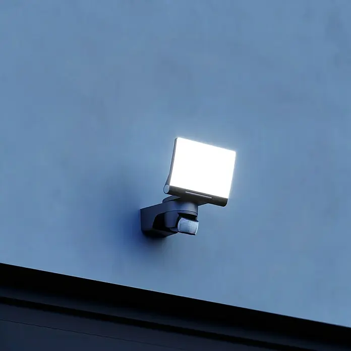 Steinel LED-Sensor-Strahler XLED home 2 S schwarz - 13,7 W, L x B x H: 161 x 180 x 218 mm, Schwarz, IP44 - Eine moderne, schwarze LED-Außenleuchte mit Bewegungsmelder ist an einer blauen Wand montiert und strahlt helles, weißes Licht aus.